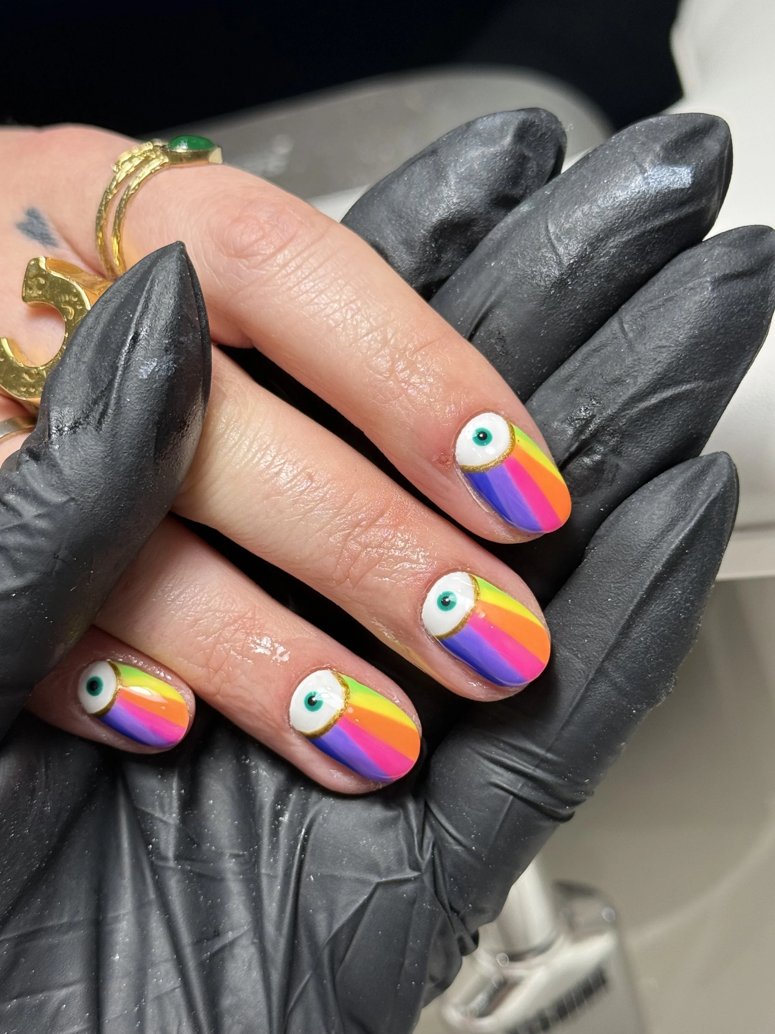 Close-up of a person's hand with rainbow-colored rainbow nail art and a rainbow eye design, wearing a gold ring with a green stone, and a black glove, possibly at a nail salon.