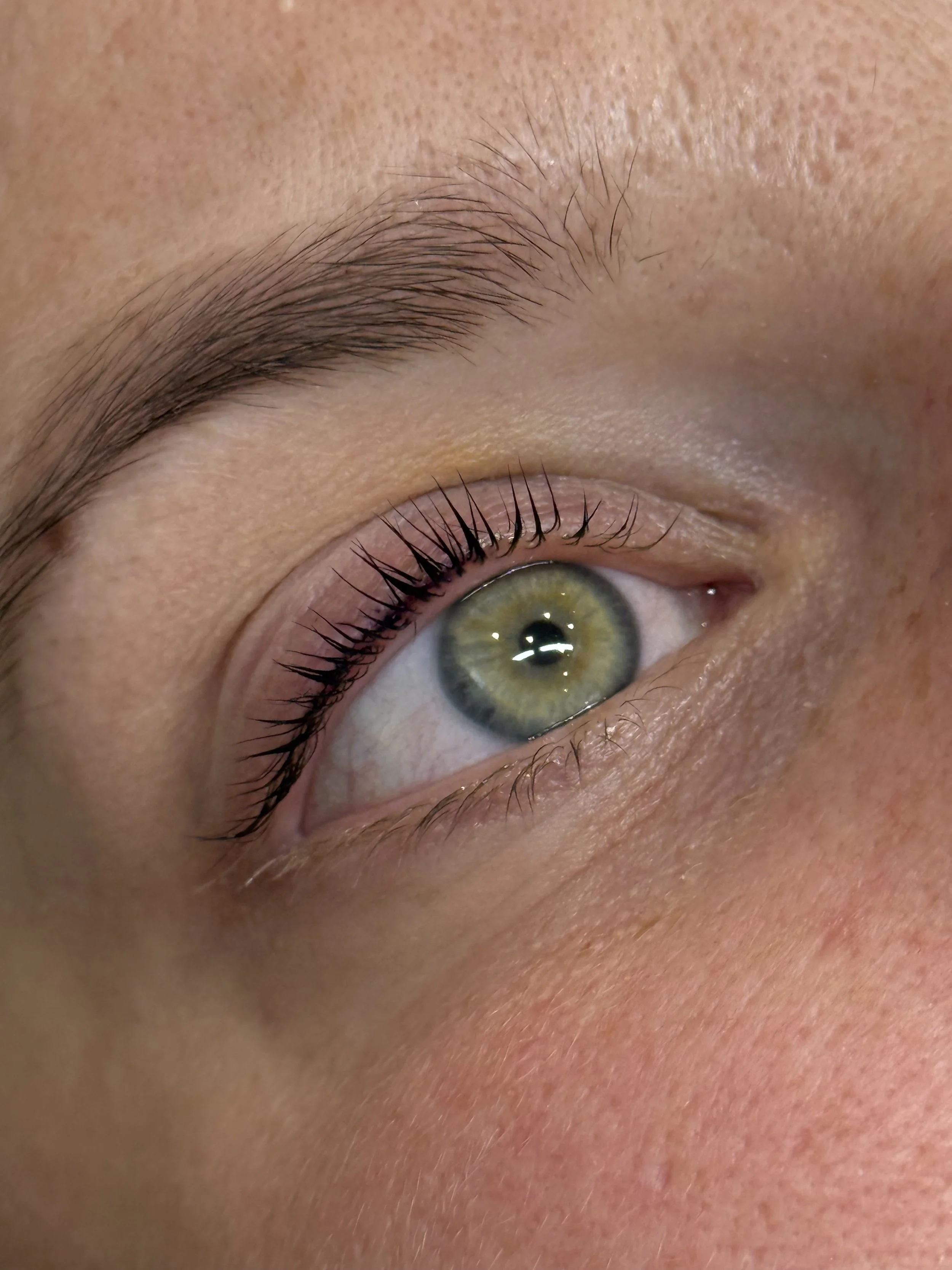 Close-up of a person's eye with green iris, long eyelashes, and well-defined eyebrow, showing detailed skin texture.
