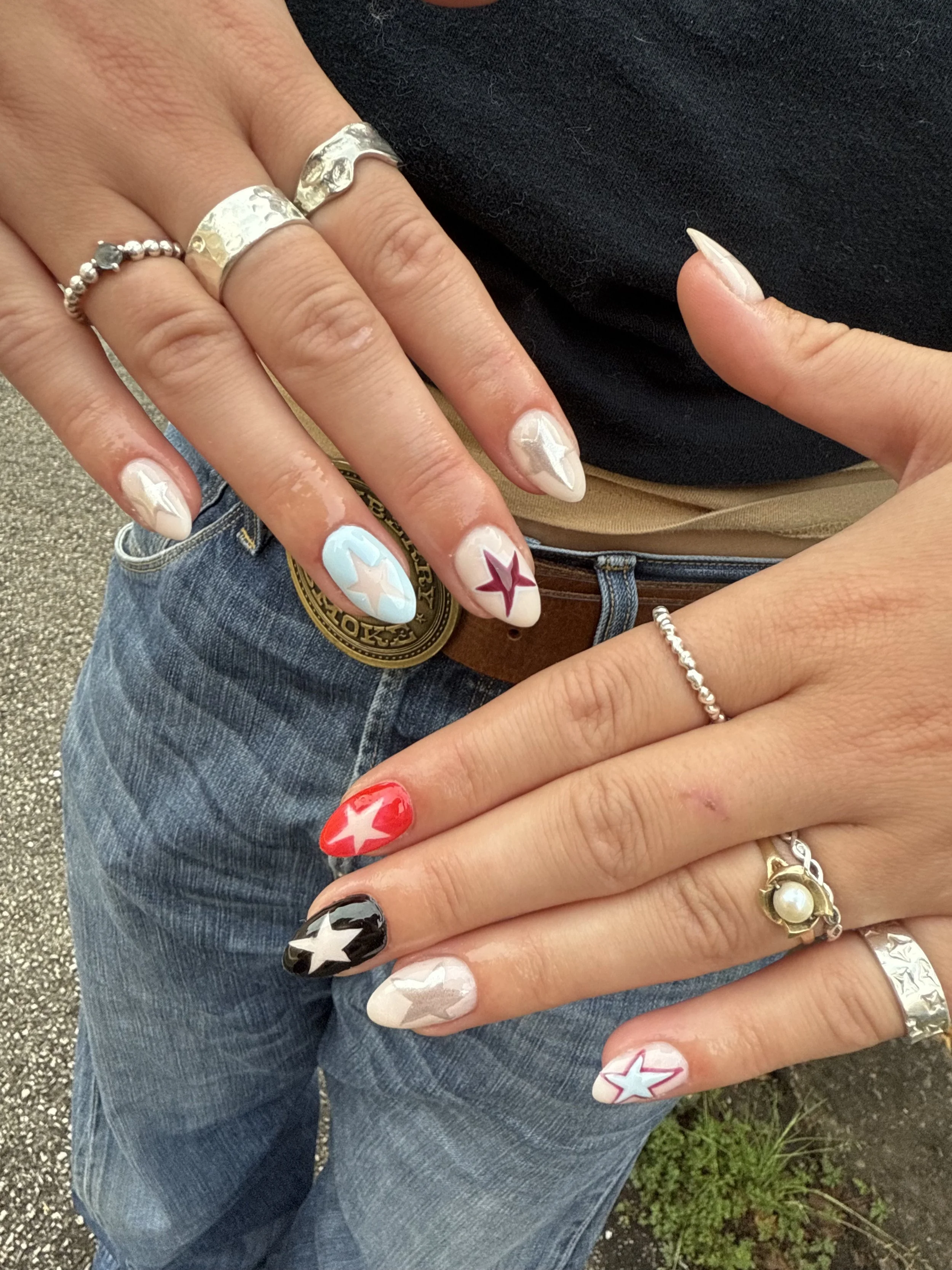 Close-up of a person's hand with decorated nails featuring stars in various colors, wearing multiple rings, including a silver hammered band, a beaded ring, and a pearl and gold ring, with part of their torso and blue jeans visible.