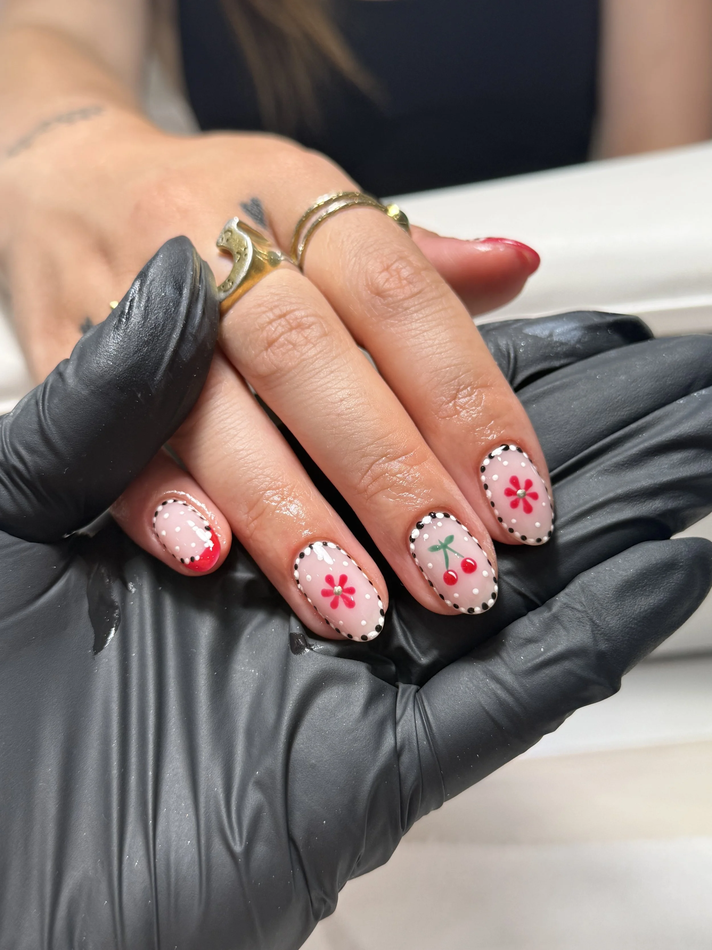 A person's hand with festive Christmas-themed nail art, including red and green cherries and red flowers, being held by a person wearing black gloves, showcasing rings on their fingers.