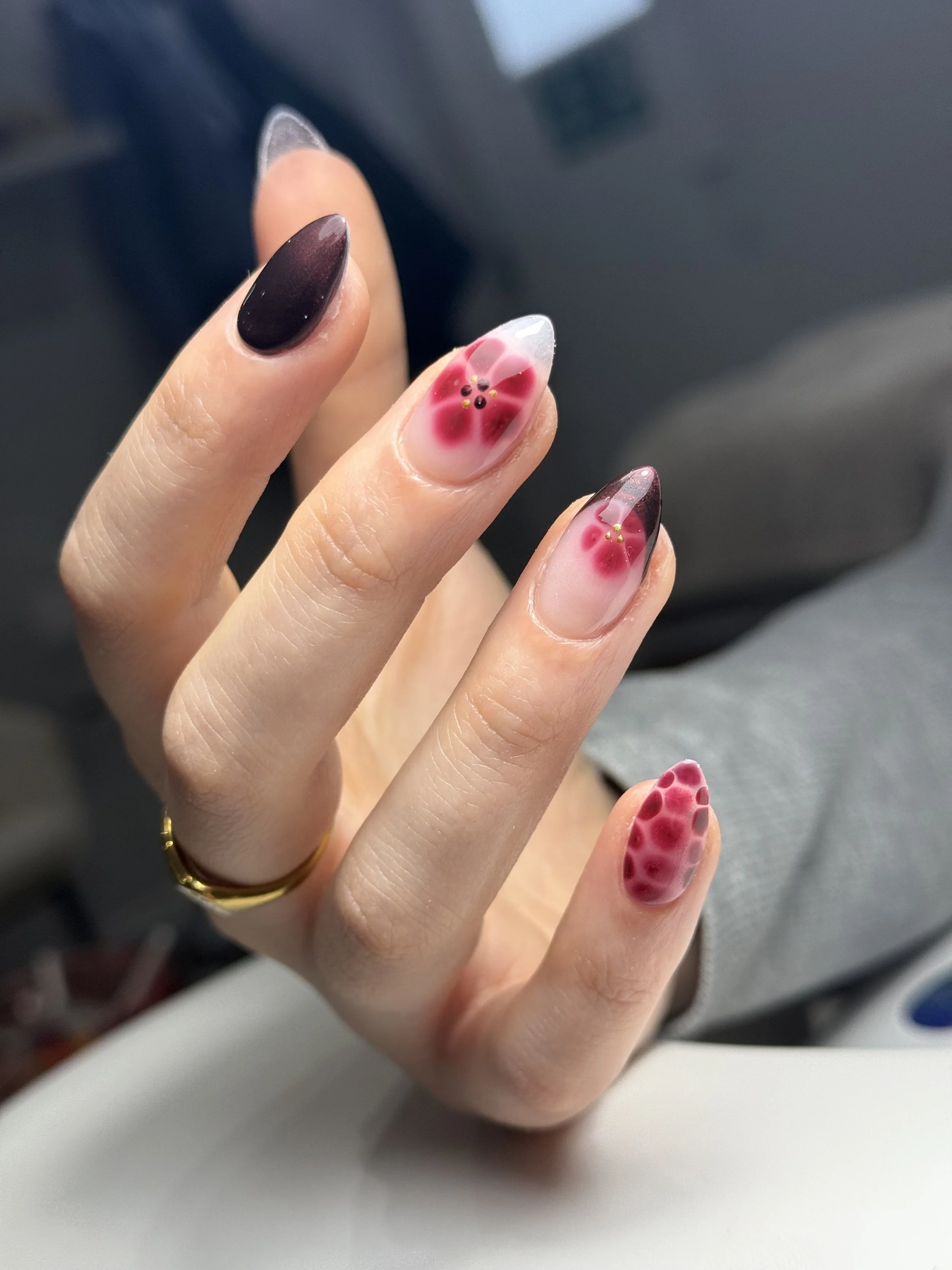 Close-up of a hand with manicured nails. The index and ring fingers feature a floral design with red flowers and black centers on a clear and pinkish base. The middle finger has a solid dark, glossy color.