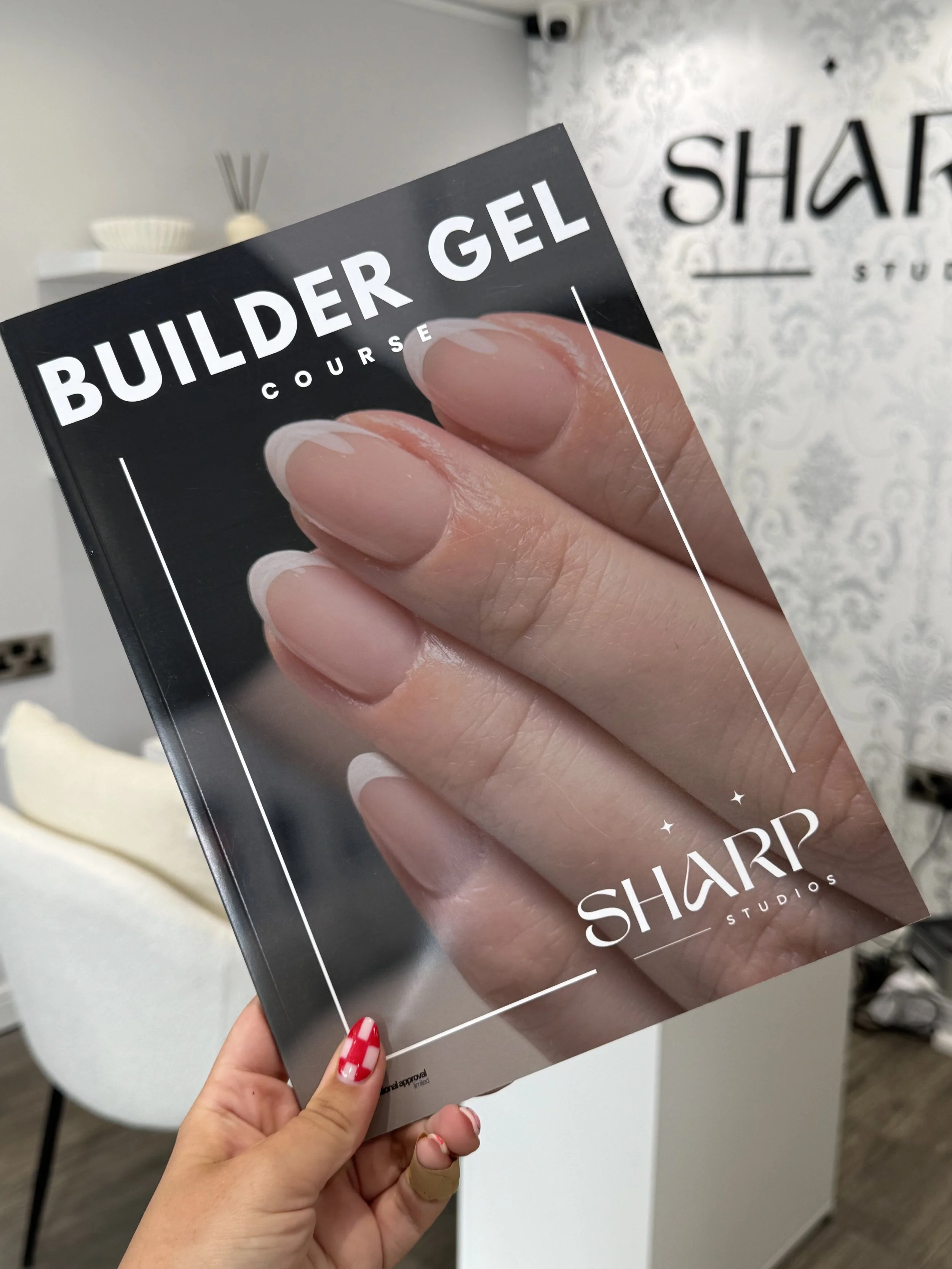 Close-up of a hand holding a Black and White pamphlet for a builder gel course at SHARP Studios, with a background of a white wall and decorative items.