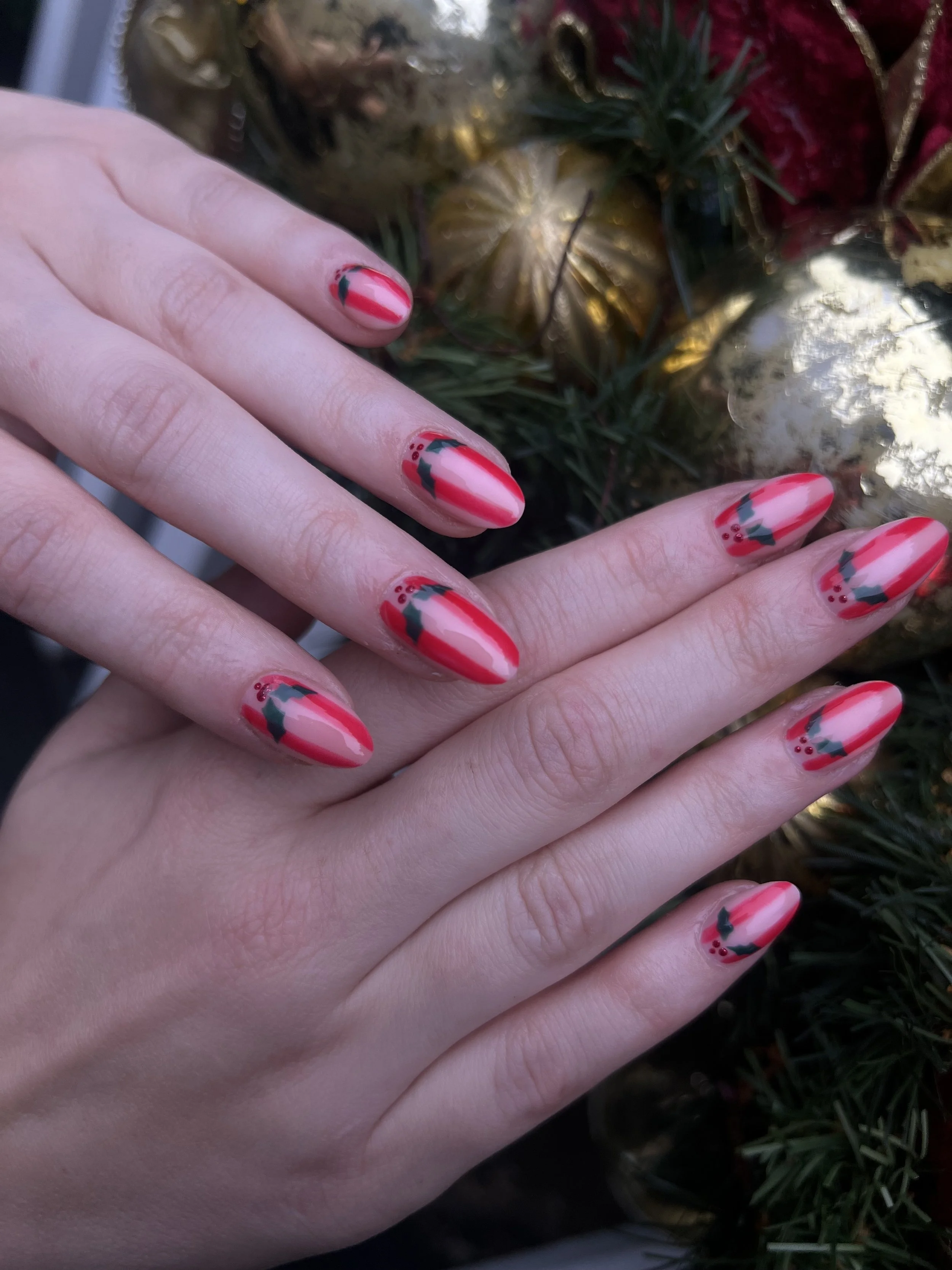 Close-up of a person's hand with festive holiday-themed nail polish, featuring red, green, and plaid patterns, against a background of Christmas ornaments and greenery.