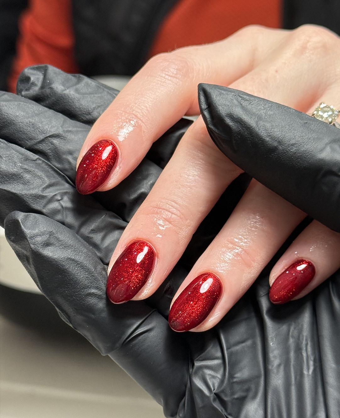 Close-up of a hand with red glittery oval-shaped nails, wearing a ring, held by a person wearing black gloves.