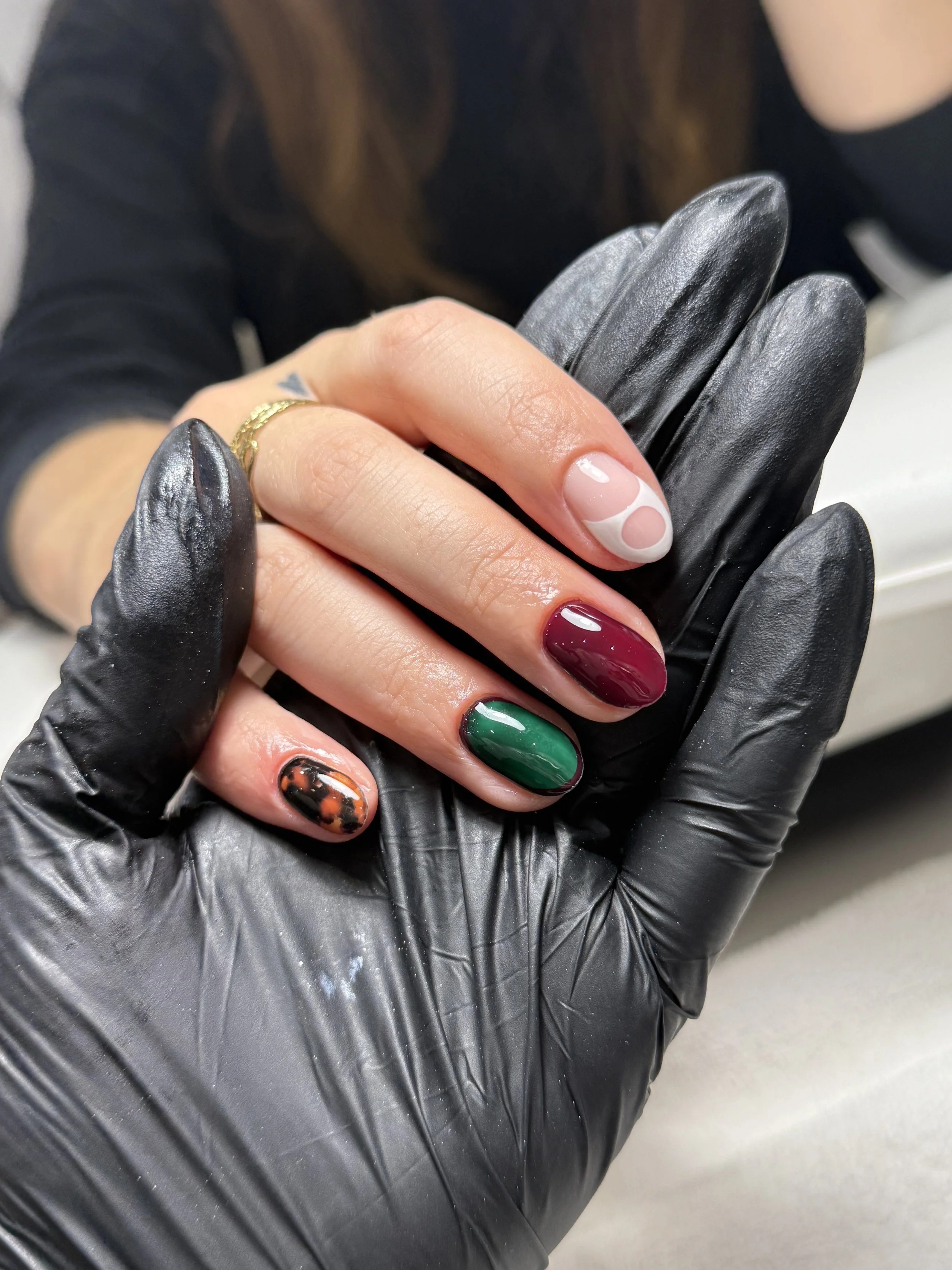 Close-up of a hand with colored nails holding a black glove, wearing a gold ring, with a person in black clothing in the background.