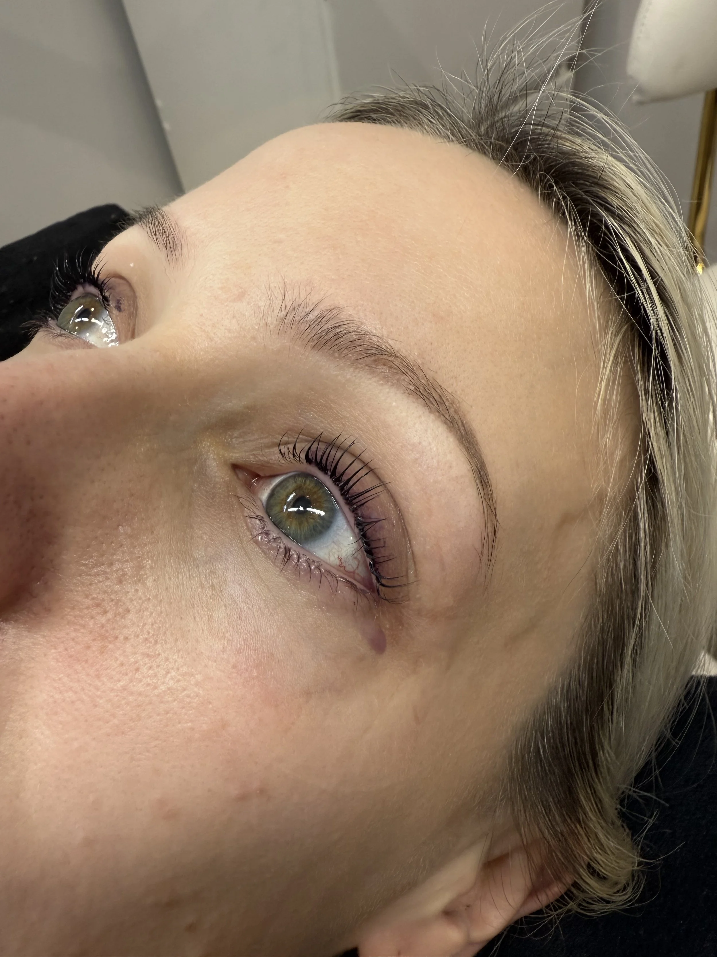 Close-up of a woman's face lying down, showing her green eyes, eyebrows, and forehead with some visible skin texture and a small mole.