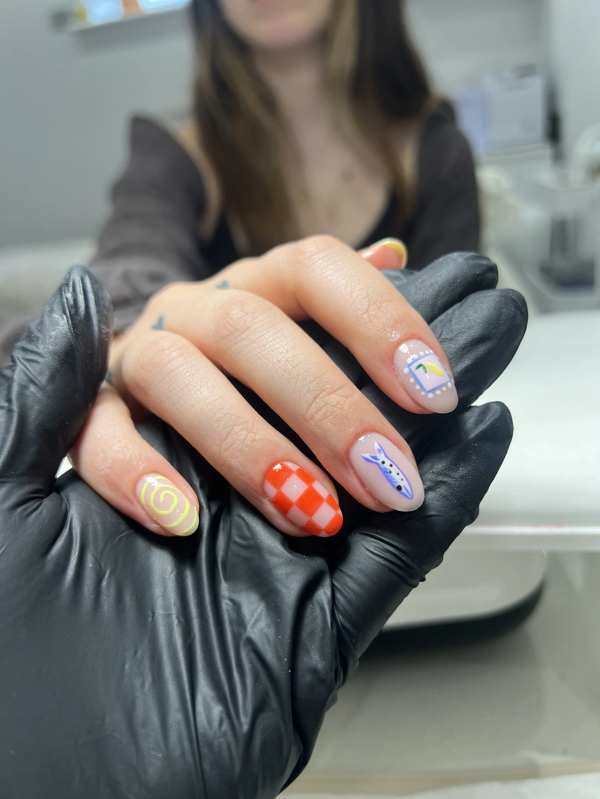 Close-up of a person's hand with decorated nails, holding another hand wearing a black glove. The nails have colorful designs including a spiral, a red and white checkered pattern, a blue animal face, and a rainbow. The background is blurred showing 