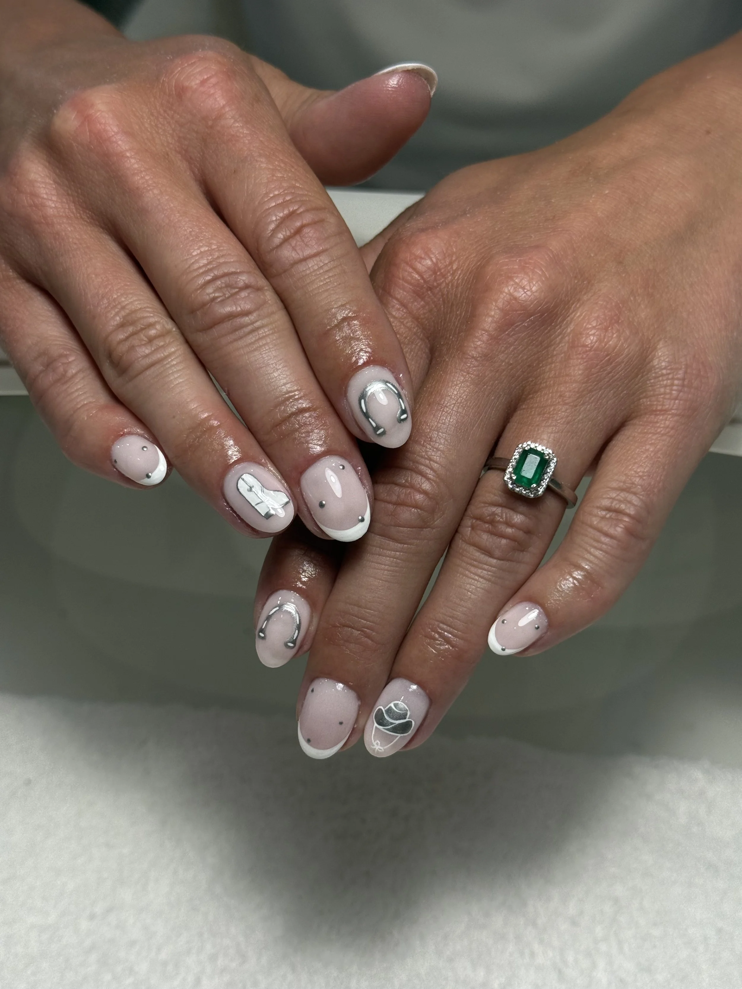 Close-up of hands with manicured nails featuring a white and gray design, including symbols like a horseshoe, a cupcake, and abstract patterns, and a ring with a large green gemstone.
