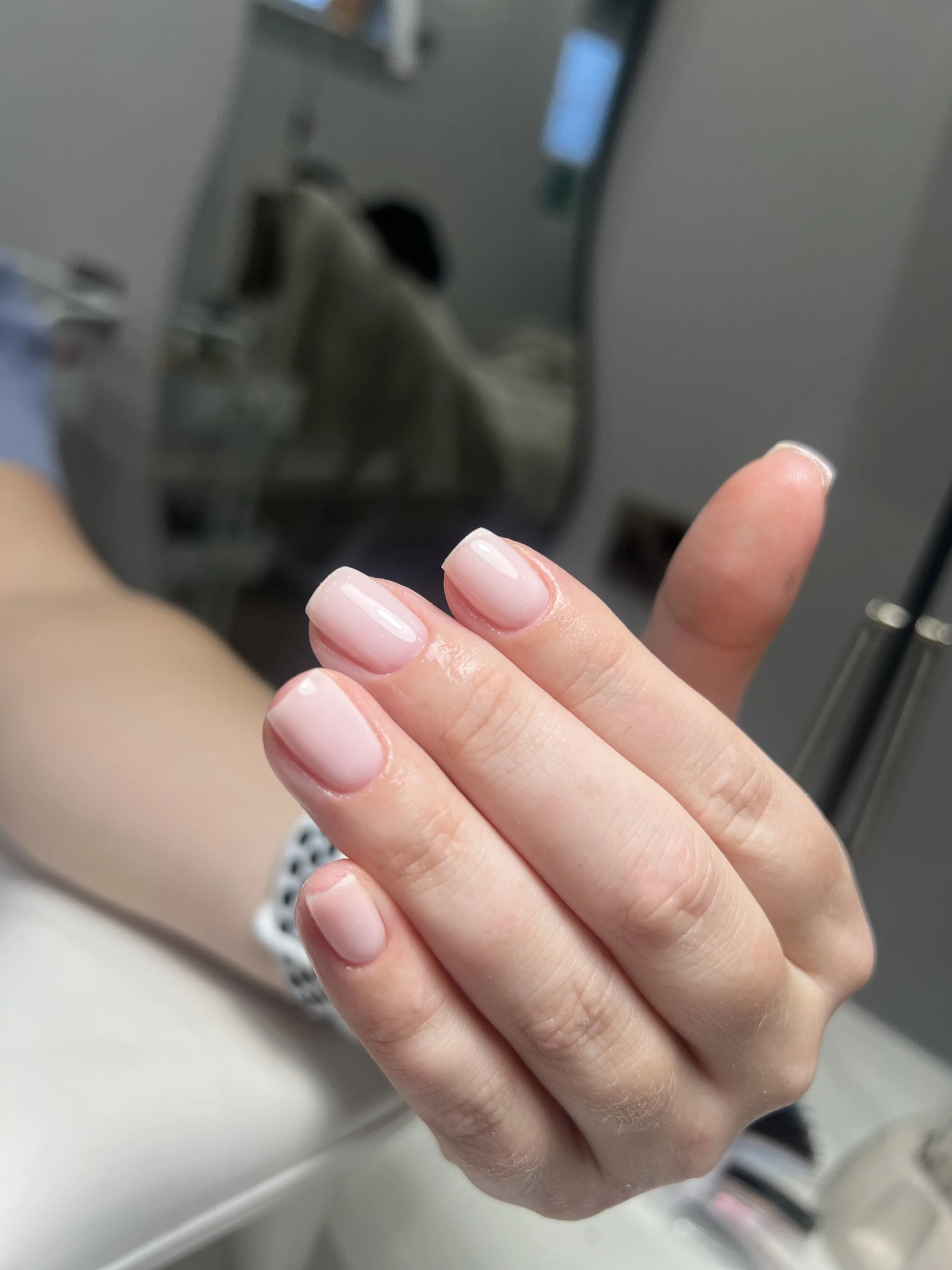 Close-up of a person's hand showing nails with a clear, shiny, and well-maintained manicure, held up against a blurred background with a mirror and medical equipment.