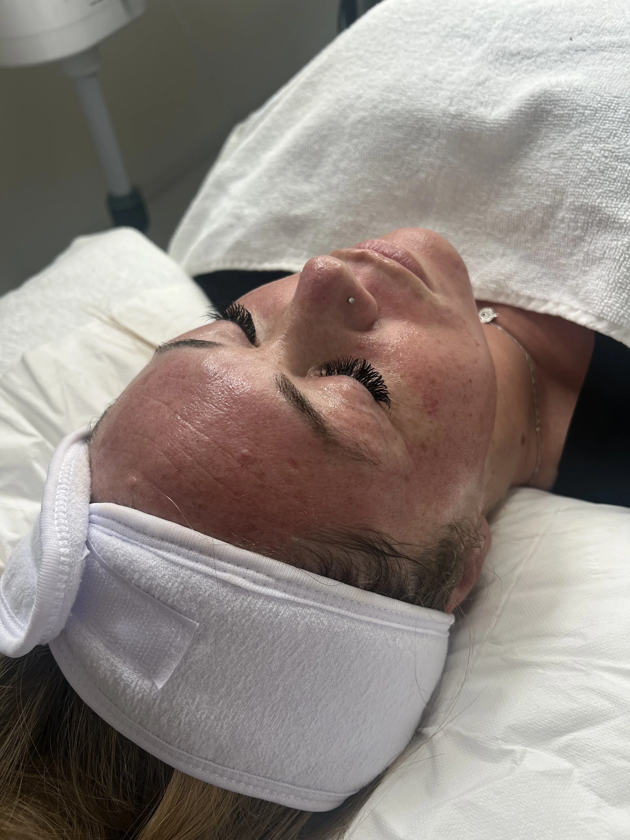 A woman lying down with her eyes closed, wearing a white headband and a white garment, appears to be receiving a facial treatment or massage.