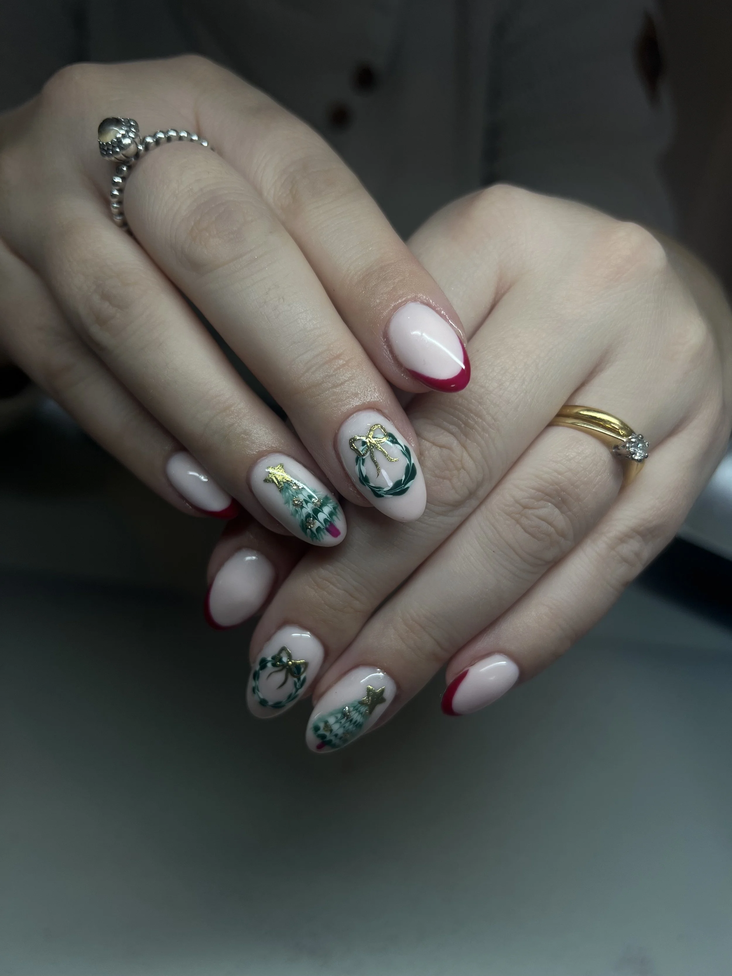 Close-up of hands showing Christmas-themed nail art with Christmas tree, wreath, and star designs, wearing a silver ball ring and a gold ring.