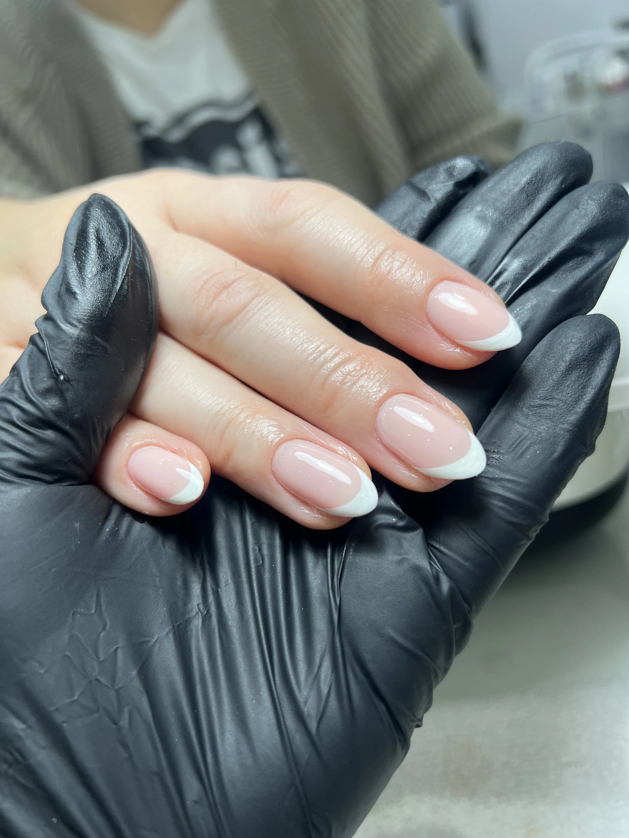 Close-up photo of a person with a manicured French nail on their hand, wearing black gloves, held by another person wearing a grey jacket.