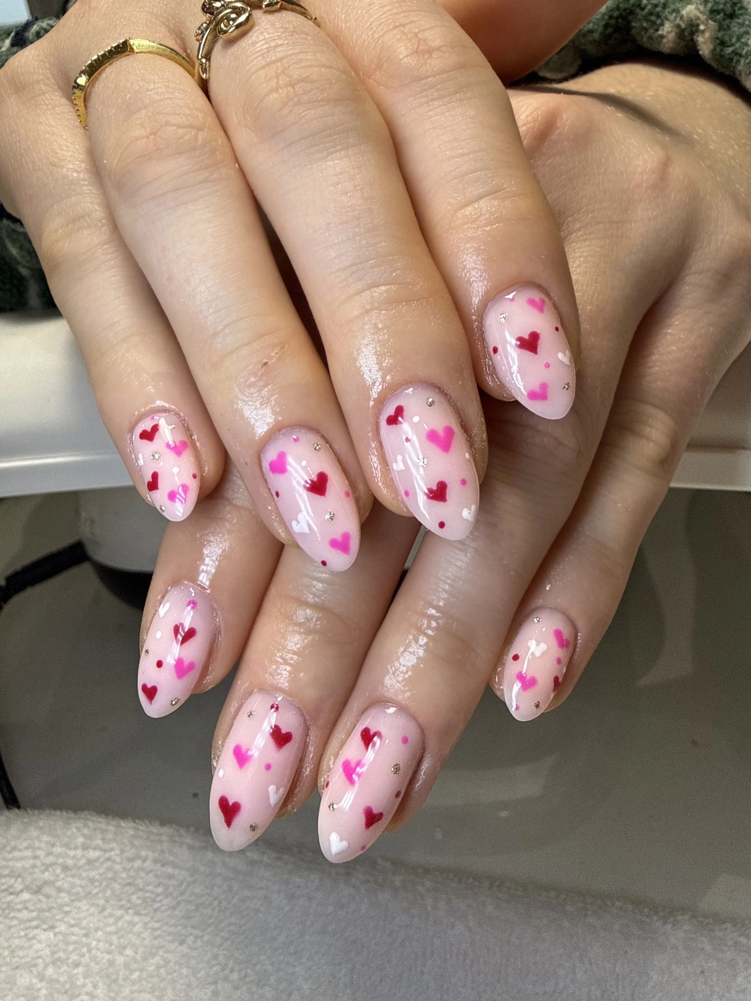 Close-up of a person's hand with pink, white, red, and purple heart-themed nail art, featuring small rhinestones. The person is wearing gold rings on their fingers.