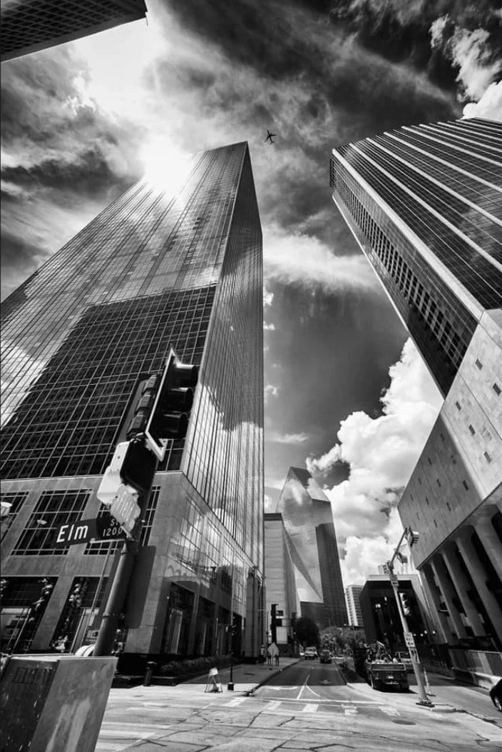 Black and white photo of tall skyscrapers in a city, with a cloud-filled sky and an airplane flying overhead.