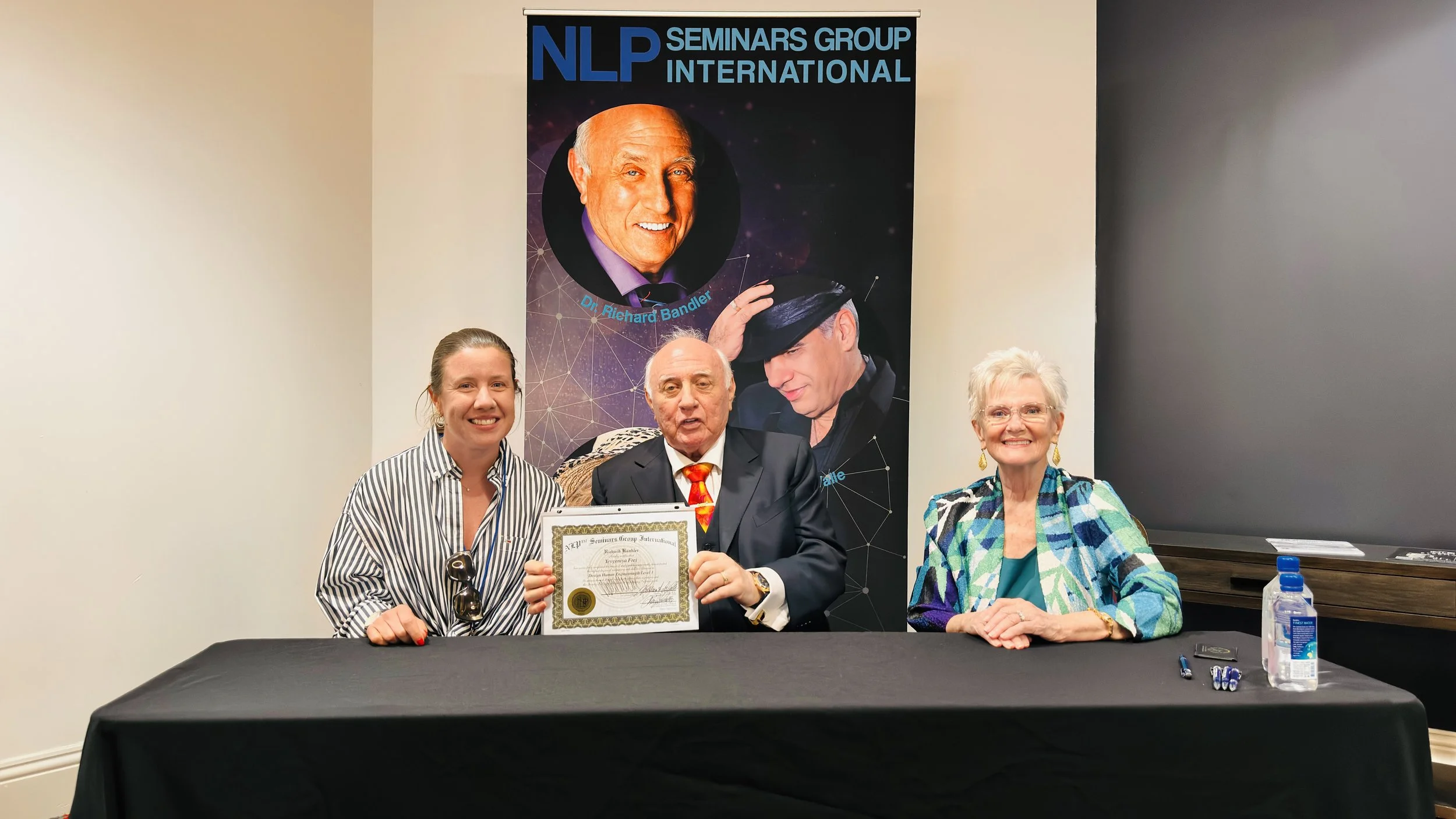 Three people sitting at a table in front of a large poster for an NLP Seminars Group International event featuring Dr. Richard Bandler. The man in the center is holding a framed certificate, flanked by two women. The table has pens, hand sanitizer, and glasses.