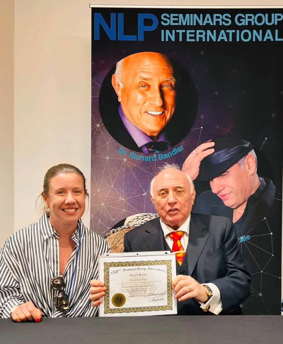 Three people sitting at a table in front of a large poster, holding a certificate. One woman on the left has sunglasses hanging from her shirt collar, and the man in the middle is wearing a suit and an orange patterned tie. The woman on the right, partially visible, is wearing a striped shirt. The poster behind them features a smiling older man with a bald head, and text reading "NLP Seminars Group International" with the name "Dr. Richard Bandler."