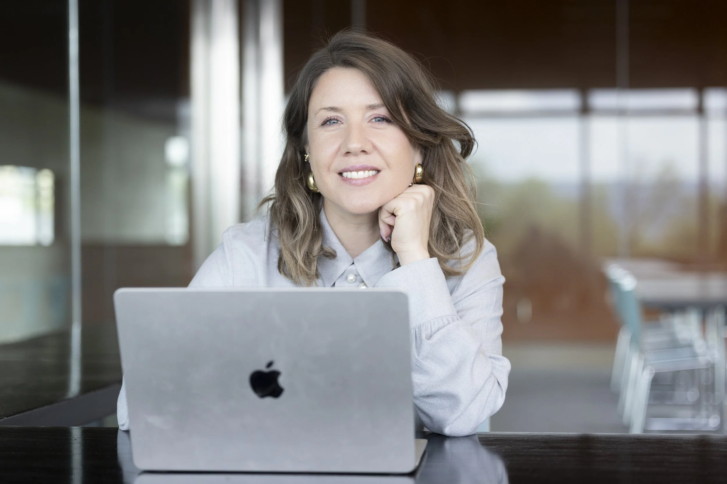 A woman with wavy brown hair and gold earrings smiling, resting her chin on her hand, sitting at a black table with an open laptop, in a bright, modern indoor setting with large windows in the background.