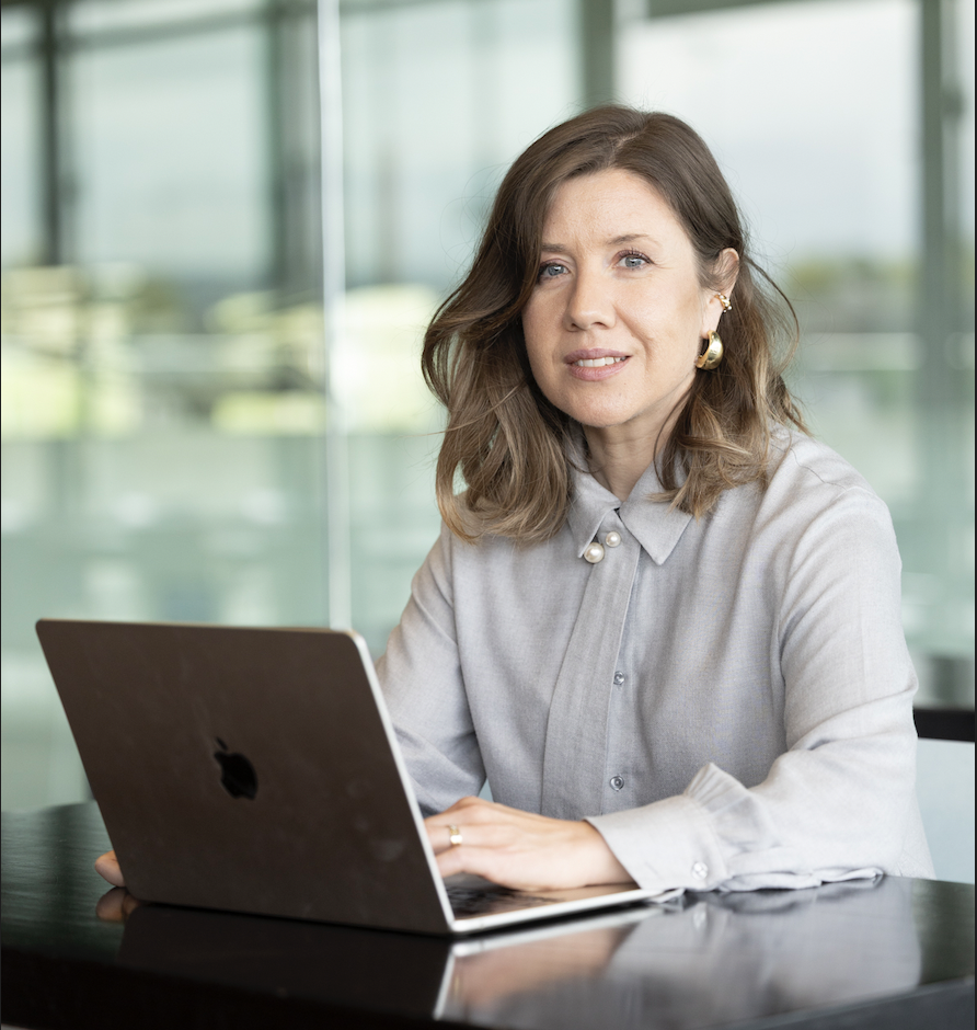 A woman with brown hair wearing a light gray blouse with pearl accents, sitting at a table with an open MacBook in front of her, in a modern office with glass windows.