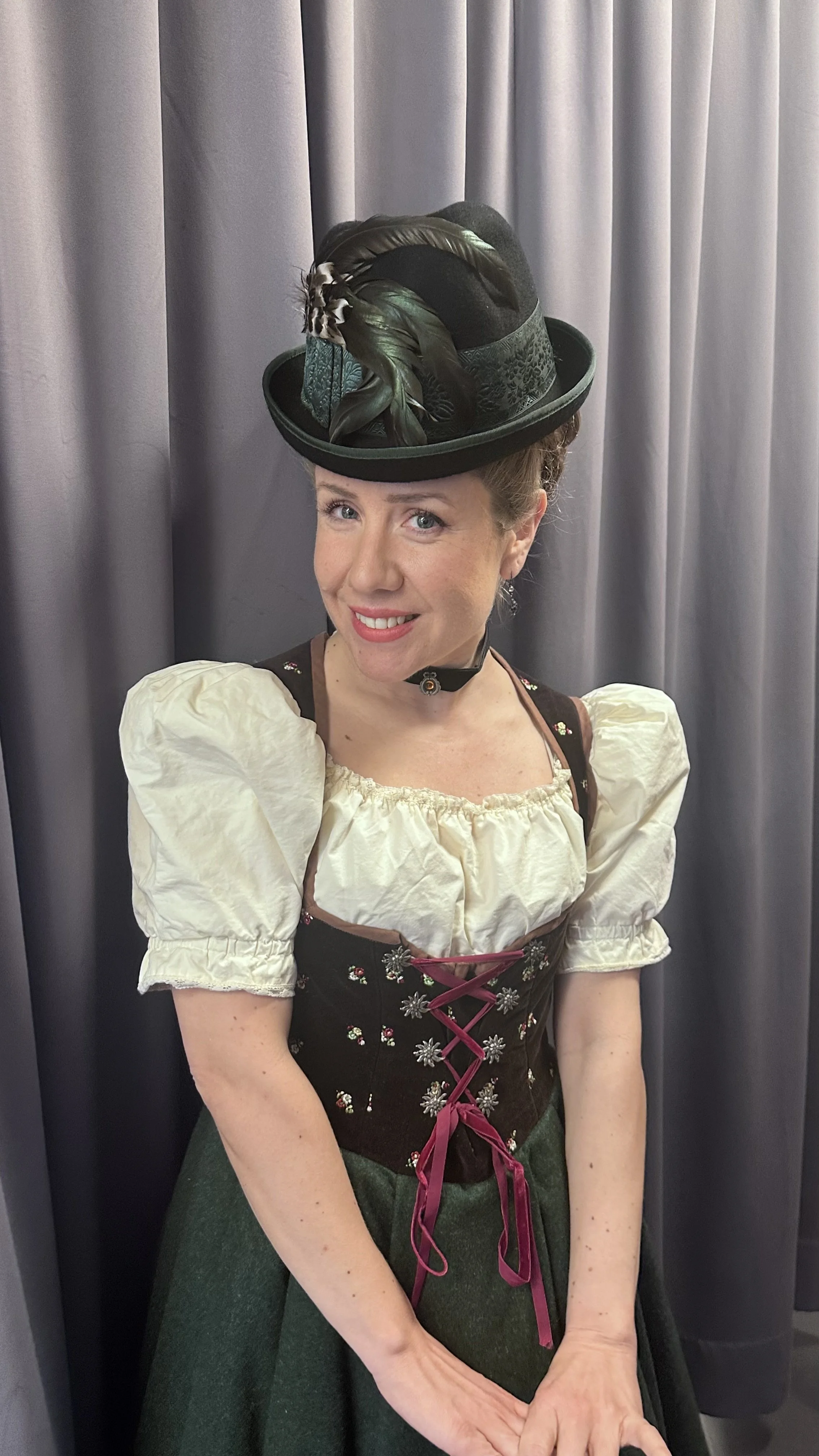A woman in traditional Bavarian dress, wearing a dirndl, with a decorative hat adorned with feathers, standing in front of a gray curtain.