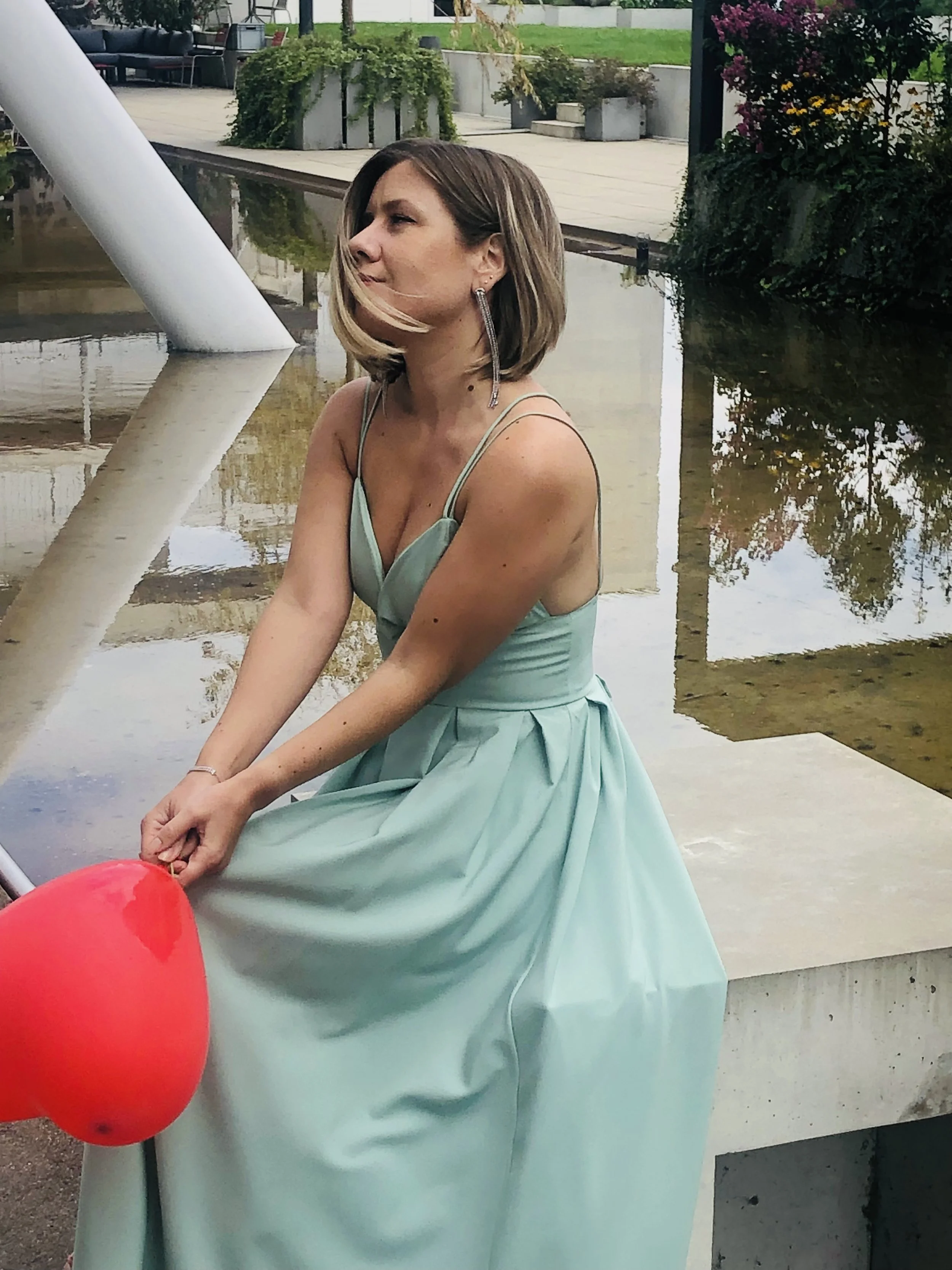 A woman with shoulder-length blond hair sitting outdoors near a water feature, wearing a mint green dress and holding a red balloon.
