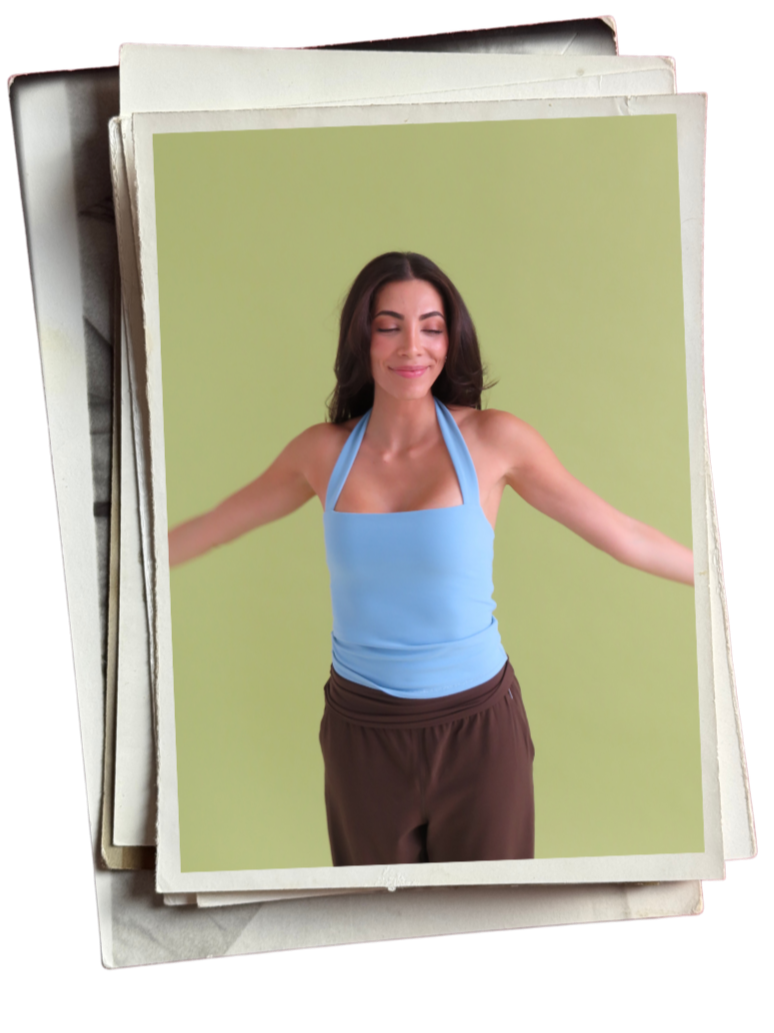 A woman with long dark hair and light skin smiling with eyes closed, wearing a blue tank top and brown pants, standing against a plain green background.