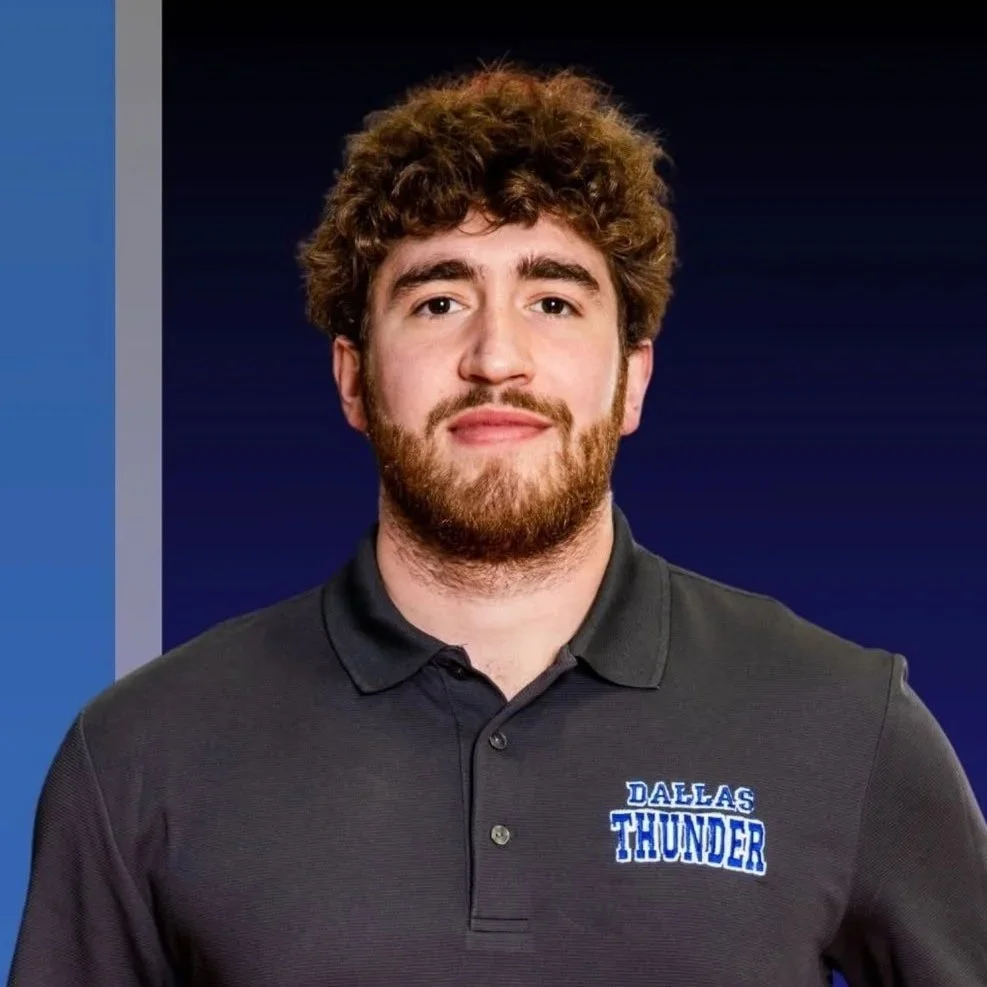 A young man with curly brown hair and a beard wearing a black polo shirt with a 'Dallas Thunder' logo, standing against a dark blue background.
