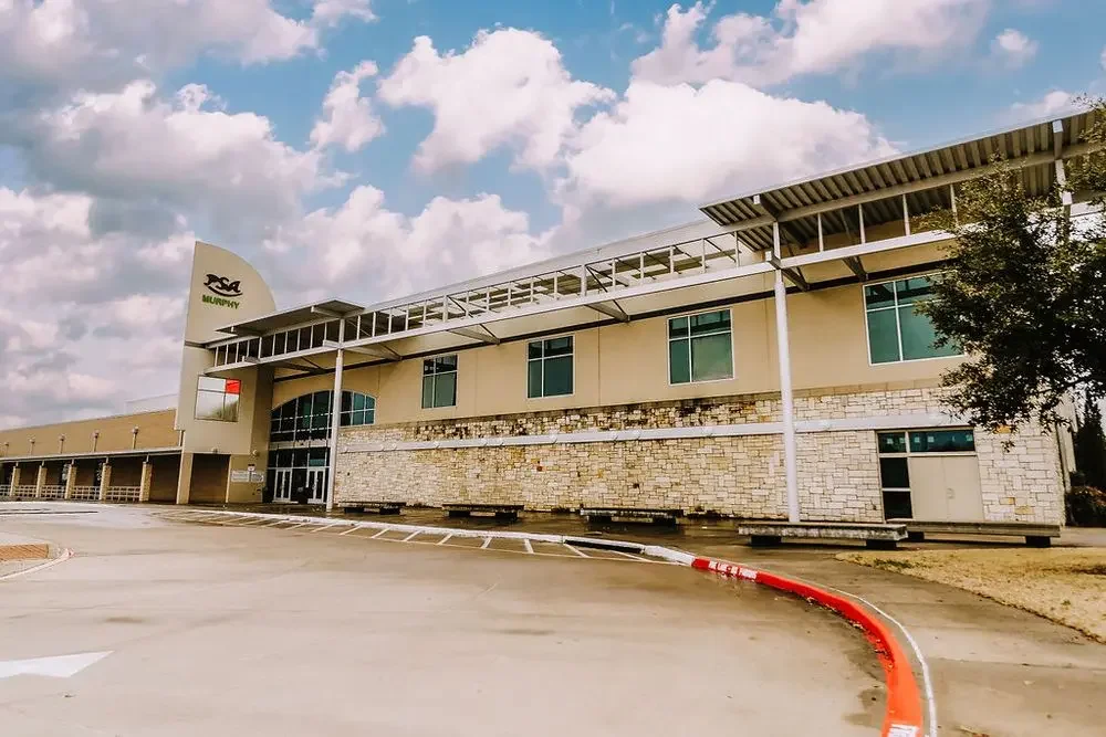 A modern commercial or office building with a stone and beige exterior, a curved row of windows, and a parking lot in front under a partly cloudy sky.