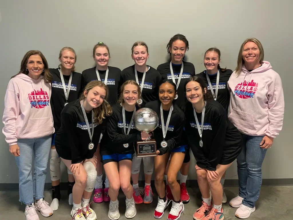 A girls' volleyball team holding a trophy, with two coaches, all smiling and wearing medals, in front of a plain gray wall.