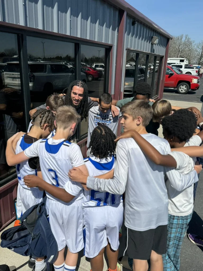 A group of young basketball players with their coach in a huddle outside a building, practicing team spirit.