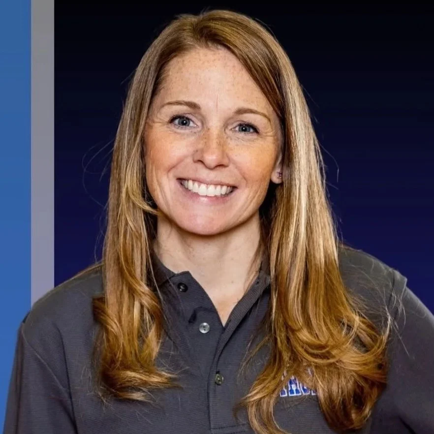 A woman with long red hair, freckles, and blue eyes smiling, wearing a dark gray collared shirt with a logo, against a dark blue background.