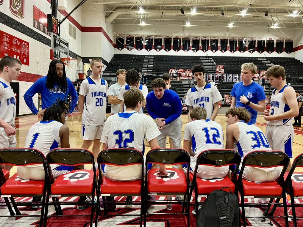 Basketball team in white and blue uniforms sitting and standing on the sidelines of a gymnasium during a timeout, listening to their coach.