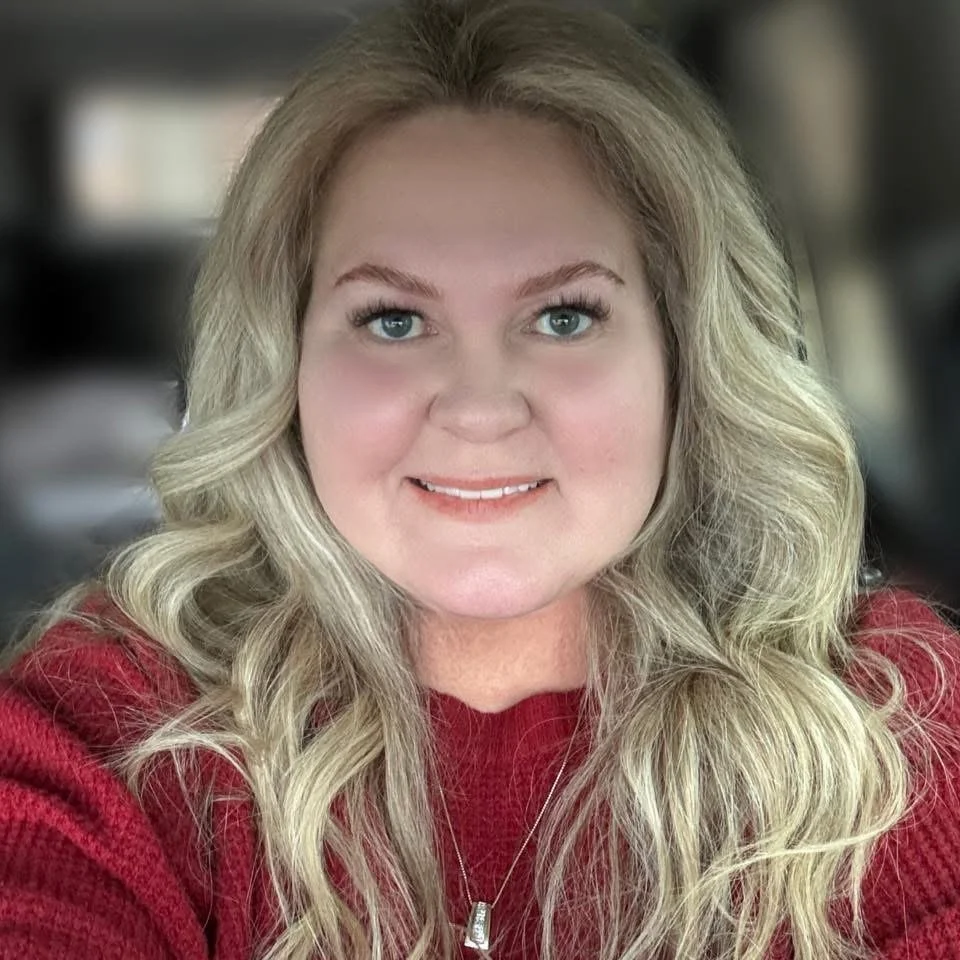 A woman with long, wavy blonde hair and blue eyes, wearing a red sweater and a silver necklace, smiling at the camera.
