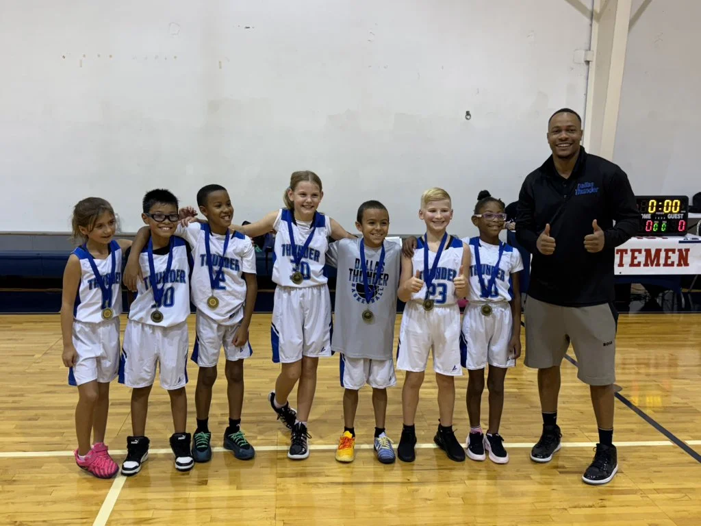 Young girls in basketball uniforms with medals, standing on a basketball court with coach giving thumbs up.