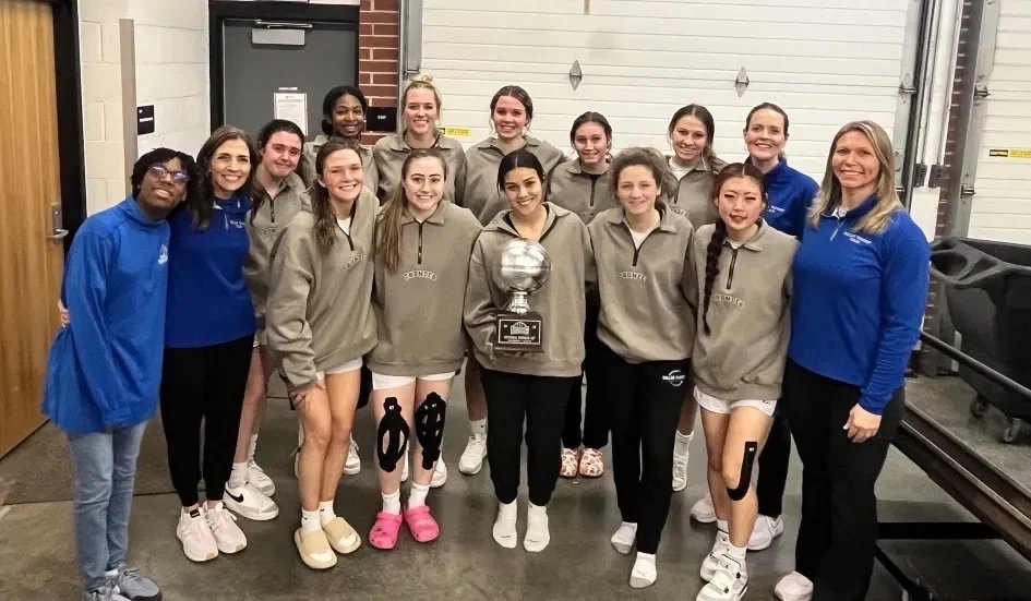 A group of women and girls posing with a trophy in a garage or storage area, some wearing tennis or sports attire.
