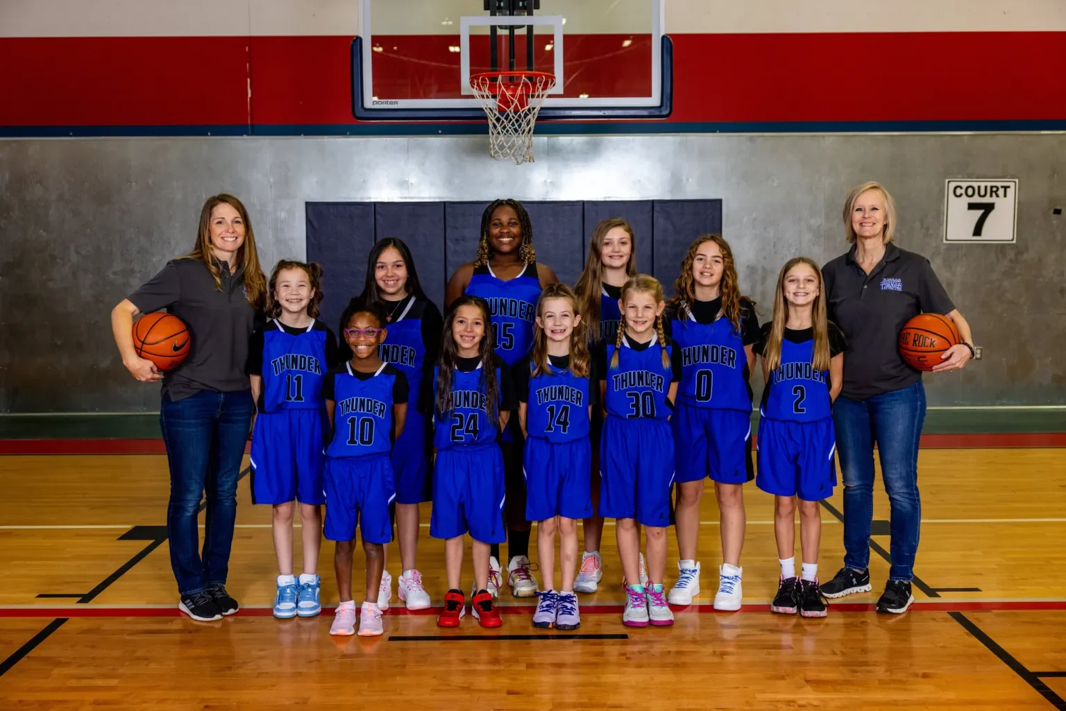 A youth girls' basketball team in matching blue uniforms with 'Thunder' and their numbers, posing in a gymnasium with two coaches holding basketballs, behind a basketball hoop.