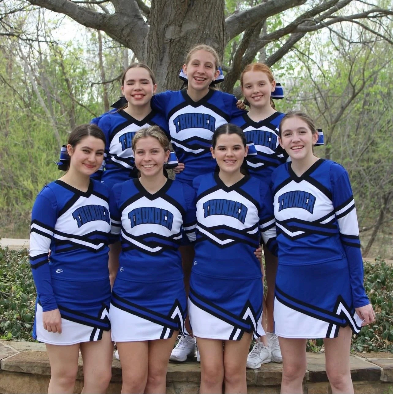 Group of young cheerleaders wearing blue and white uniforms with the word 'Thunder' on the front, standing outdoors in front of a large tree, smiling for a photo.
