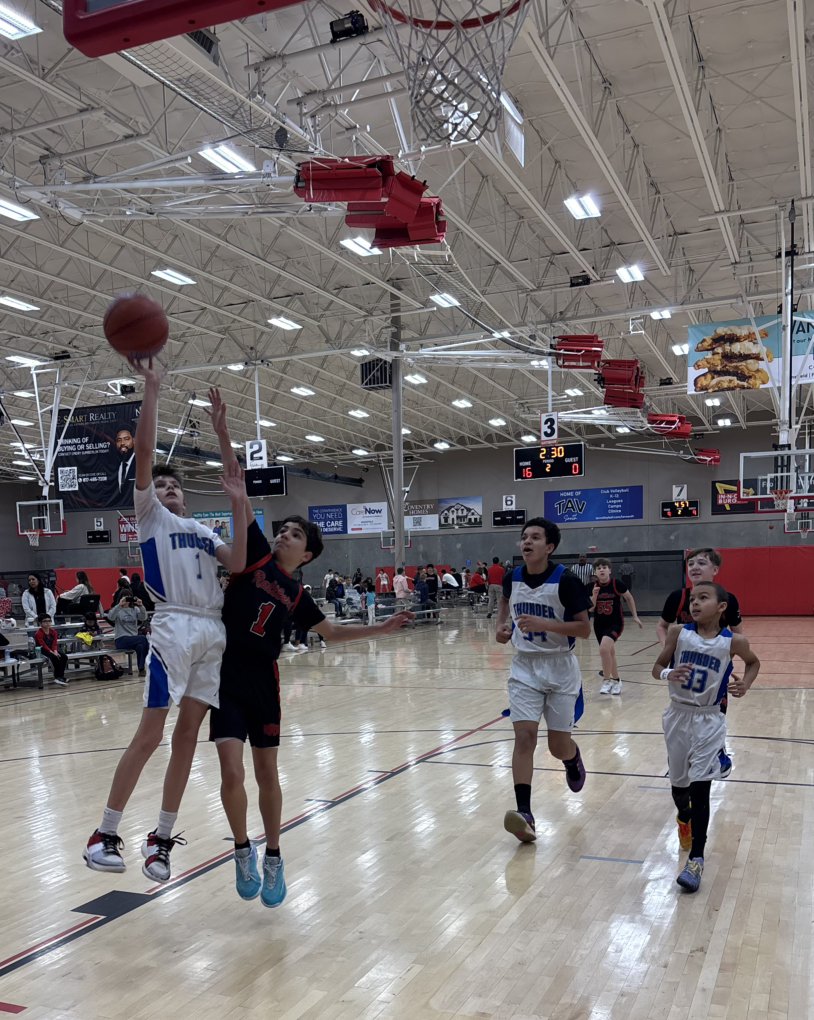 Boys playing basketball in an indoor gym, with some children wearing uniforms that say 'Thunder' and others in black uniforms, with basketball hoops and scoreboard overhead.