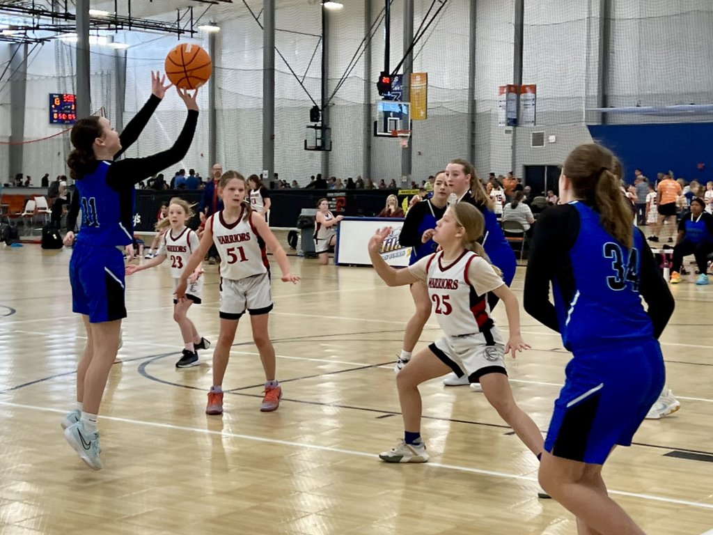 Young girls playing basketball in an indoor gym, with one girl in blue jumping to shoot the ball, and other girls in white and blue waiting or defending.