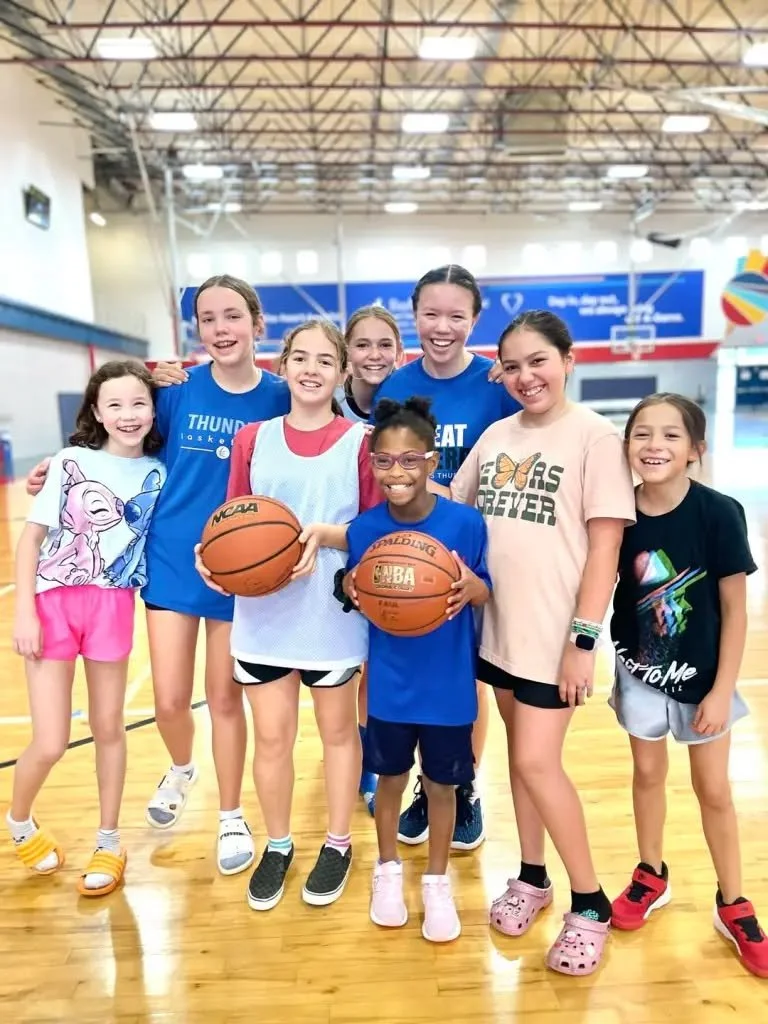 Group of young girls in a gymnasium, some holding basketballs, posing for a photo, smiling.