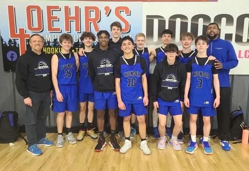 Group of young basketball players in blue uniforms with their coaches, standing on a wooden court in front of a large banner at an indoor gym.