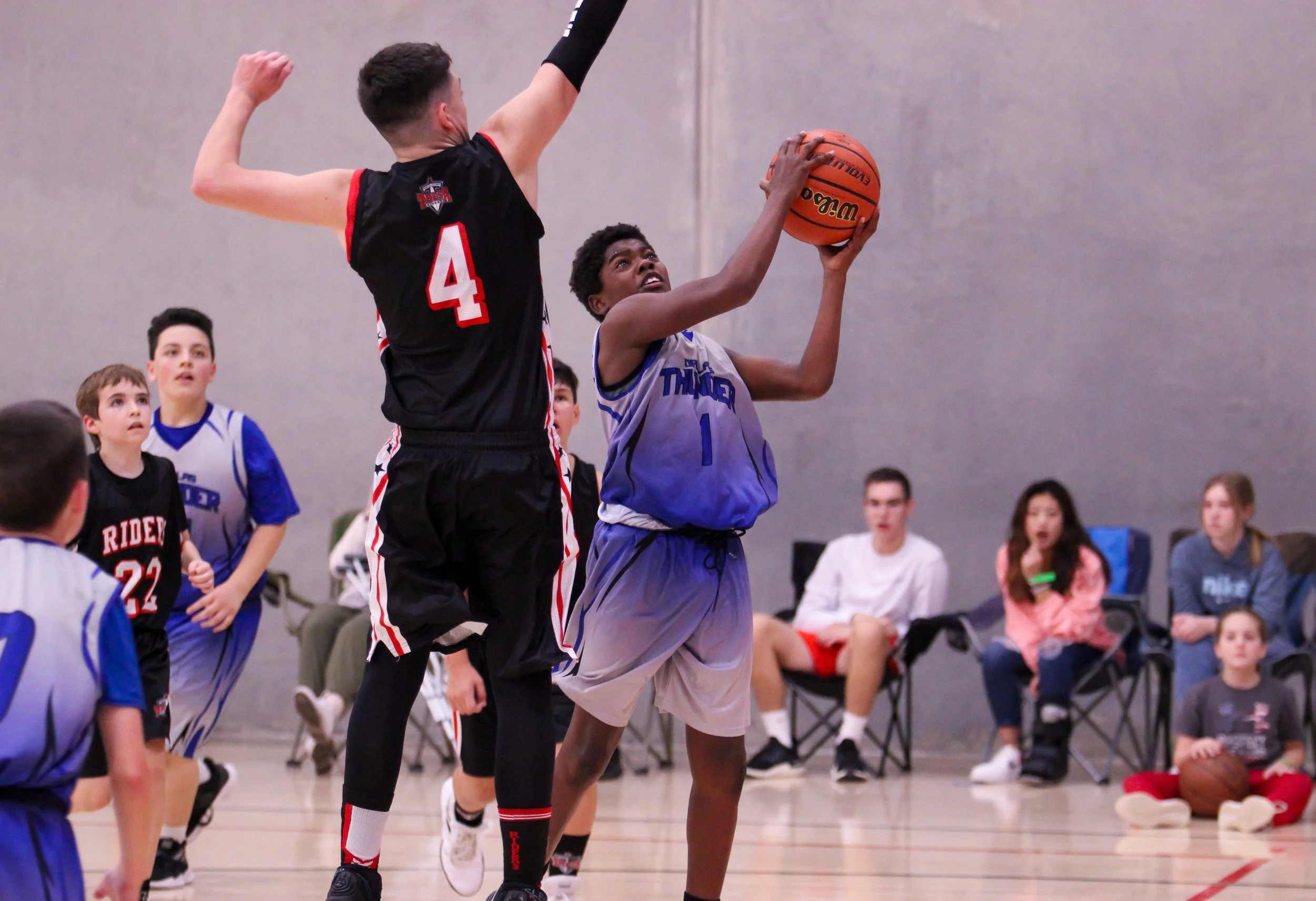 A group of young boys playing basketball indoors, with one boy in a blue jersey about to shoot the ball while a taller player in a black jersey with the number 4 attempts to block the shot. Spectators are watching in the background.
