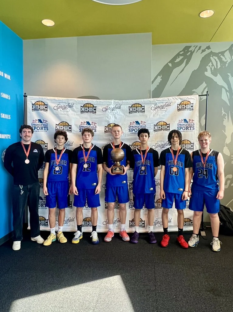 Group of six young basketball players in Blue Thunder jerseys and their coach standing in front of a backdrop with sports logos. The team is celebrating a victory, with medals around their necks and one player holding a trophy.