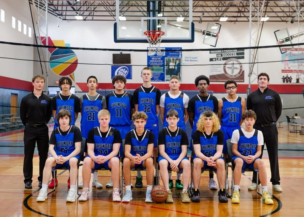 A high school basketball team called the Thunder, consisting of ten players and two coaches, poses for a team photo in a gymnasium. The players are wearing blue jerseys with black and white accents, and the coaches are in black attire. The gym has a basketball hoop, colorful wall mural, and banners hanging in the background.