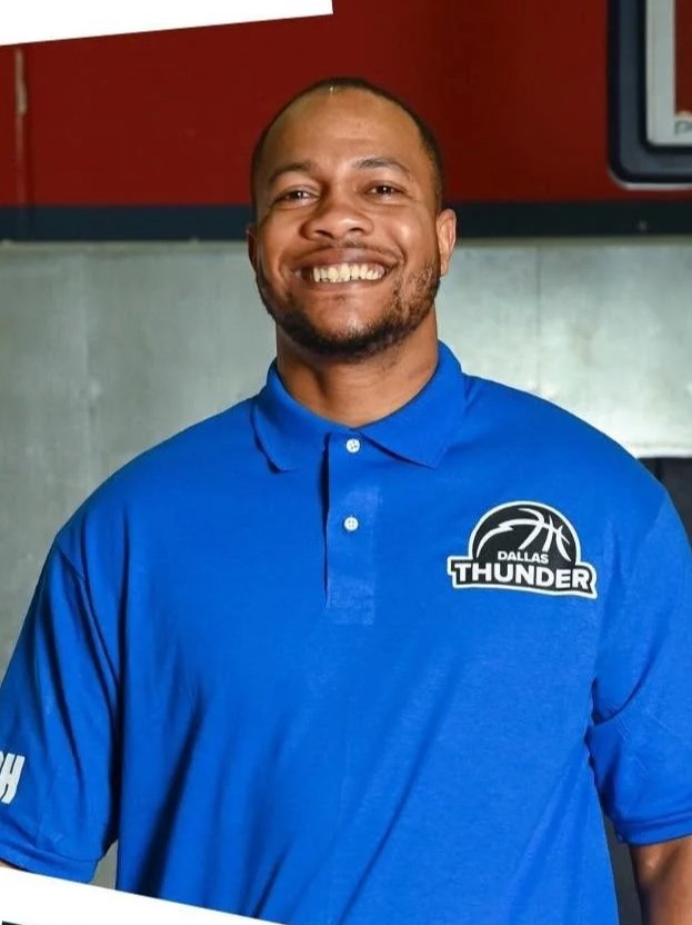 A smiling man wearing a blue Dallas Thunder sports polo shirt with a basketball logo, standing indoors in front of a red wall.
