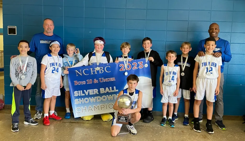 Youth basketball team celebrating with medals and a large plaque after winning the 2023 Silver Ball Showdown Championship; team members and coaches pose in front of a blue wall, holding a banner and a trophy.