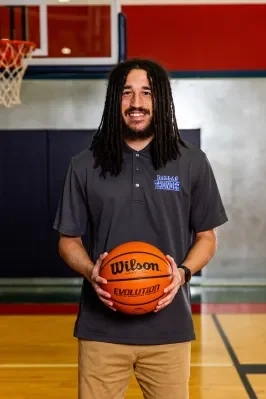 Man with long dreadlocks smiling and holding a basketball on a basketball court, wearing a dark polo shirt and khaki pants.