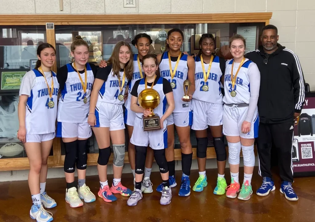 A girls' basketball team in white and blue uniforms posing with a coach, holding trophies and medals, after a game or tournament.