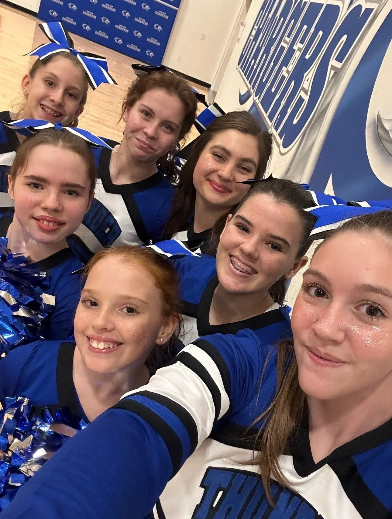 Group of young cheerleaders wearing blue and white uniforms taking a selfie in front of a sports team poster.