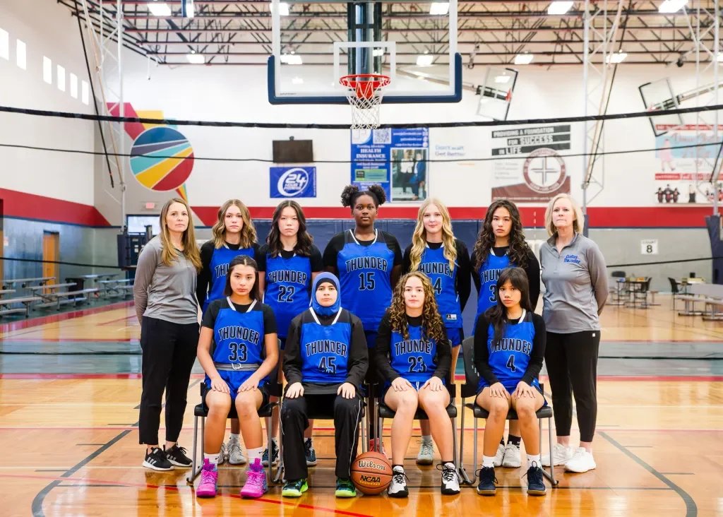 Girls' basketball team in blue and black uniforms with coaches, posing for a team photo inside a gymnasium with a basketball hoop and court.