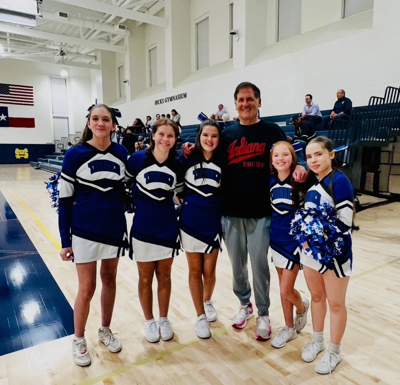A group of five young female cheerleaders in blue and white uniforms with pom-poms, standing with a male coach in a gymnasium. The background shows bleachers, an American flag, and the sign 'Hicks Gymnasium'.